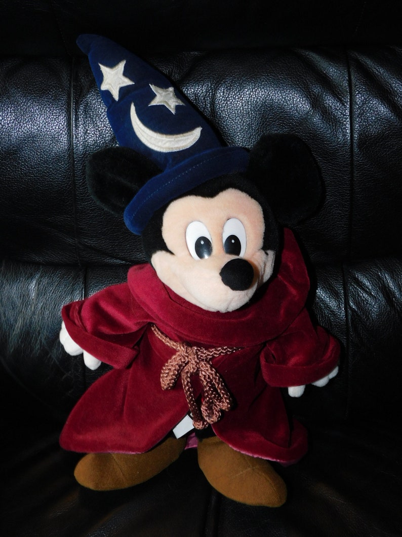 May include: A plush Mickey Mouse dressed as a sorcerer, wearing a dark blue hat with white stars and a crescent moon. He has a burgundy velvet robe and brown shoes. The toy is on a black leather surface.