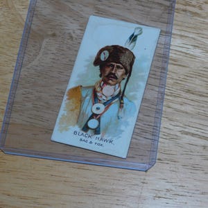 May include: A vintage trading card featuring a portrait of Black Hawk, Sac & Fox. The card shows a man in a fur hat with a feather, a light blue jacket, and a decorative necklace. The card is in a clear plastic sleeve.