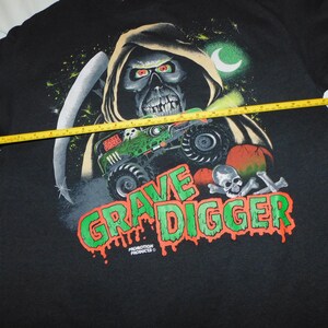 Może przedstawiać: Czarny t-shirt z grafiką Grim Reaper, monster truckiem i napisem "Grave Digger" w kolorze zielonym i pomarańczowym. Ż&oacute;łta miarka służy do pomiaru koszulki.