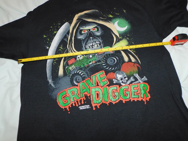 Może przedstawiać: Czarny t-shirt z grafiką Grim Reaper, monster truckiem i napisem "Grave Digger" w kolorze zielonym i pomarańczowym. Ż&oacute;łta miarka służy do pomiaru koszulki.