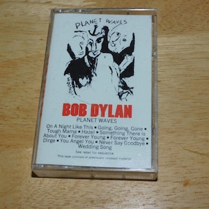 May include: A vintage cassette tape of Bob Dylan's "Planet Waves" album. The cover features a black and white illustration of several faces and the album title in red. The tracklist is visible on the bottom.