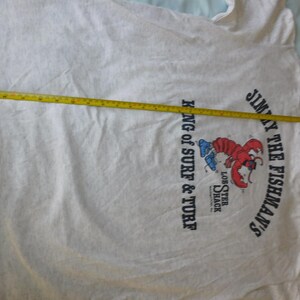 Op de afbeelding: Een lichtgrijs t-shirt met een afbeelding van een rode kreeft en de tekst "JIMMY THE FISHMAN'S" en "KING OF SURF & TURF". Een gele rolmaat ligt over het shirt en meet de lengte in centimeters.