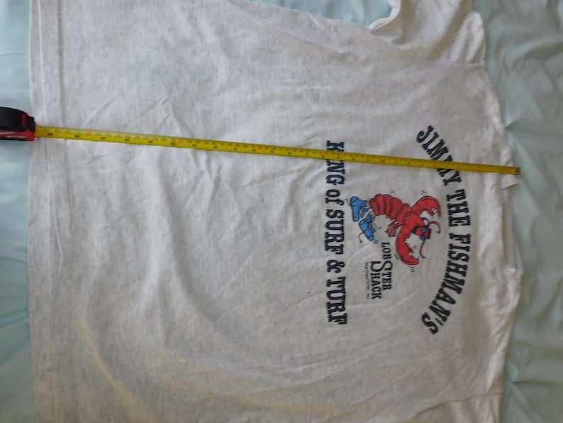 Op de afbeelding: Een lichtgrijs t-shirt met een afbeelding van een rode kreeft en de tekst "JIMMY THE FISHMAN'S" en "KING OF SURF & TURF". Een gele rolmaat ligt over het shirt en meet de lengte in centimeters.