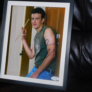 May include: A framed photograph of a person with dark hair wearing a green sleeveless shirt and blue jeans, holding a wooden drumstick. The image features a blue ink autograph across the center and a small authentication sticker in the bottom corner.