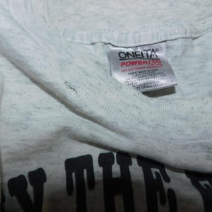 Op de afbeelding: Close-up van een lichtgrijs t-shirt met een zichtbaar label. Het label leest "ONEITA POWER/55" in rode en zwarte tekst. Het shirt heeft een licht versleten uiterlijk.