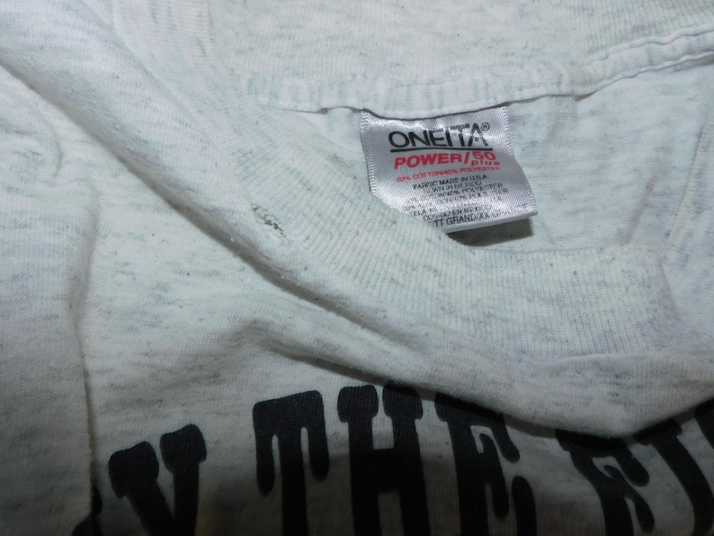 Op de afbeelding: Close-up van een lichtgrijs t-shirt met een zichtbaar label. Het label leest "ONEITA POWER/55" in rode en zwarte tekst. Het shirt heeft een licht versleten uiterlijk.
