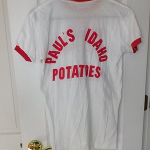 May include: White t-shirt with red trim on the collar and sleeves. The back of the shirt features the words "Paul's Idaho Potatoes" in red, arched across the top and bottom of the shirt. The shirt is hanging on a black hanger.