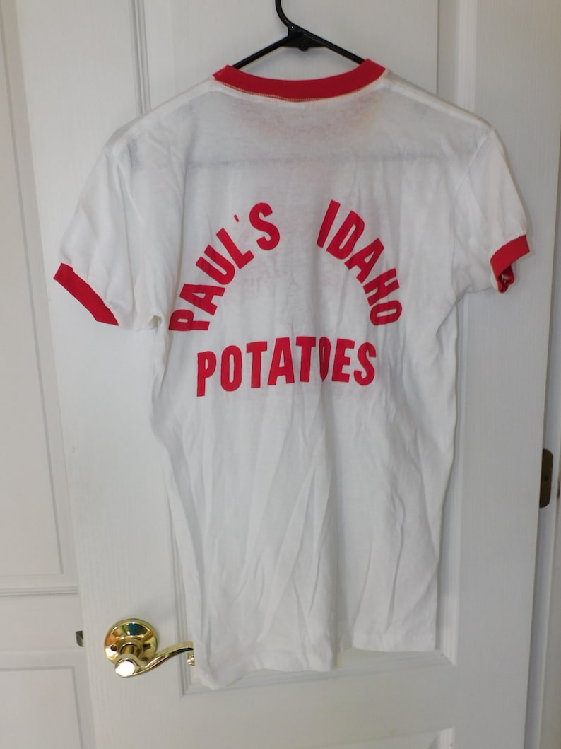May include: White t-shirt with red trim on the collar and sleeves. The back of the shirt features the words "Paul's Idaho Potatoes" in red, arched across the top and bottom of the shirt. The shirt is hanging on a black hanger.