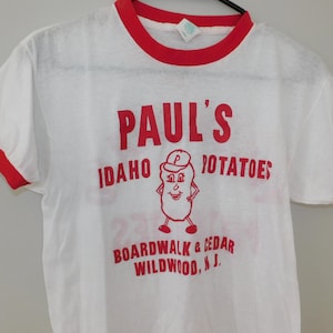 May include: White t-shirt with red trim featuring the text "PAUL'S IDAHO POTATOES" in red, along with a cartoon potato character. The shirt also includes the text "BOARDWALK & CEDAR WILDWOOD, N.J."