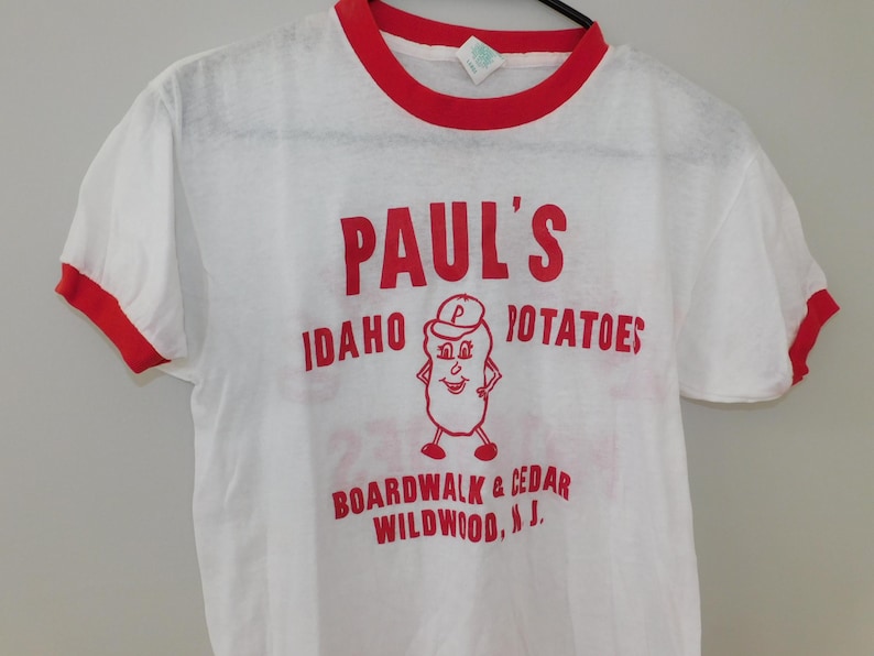 May include: White t-shirt with red trim featuring the text "PAUL'S IDAHO POTATOES" in red, along with a cartoon potato character. The shirt also includes the text "BOARDWALK & CEDAR WILDWOOD, N.J."