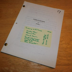 May include: A script page titled "Picking Up The Pieces" by Bill Wilson, with a yellow sticky note listing names. The page is held together with brass fasteners and rests on a wooden surface.