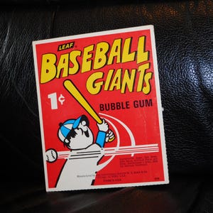 May include: A rectangular Leaf Baseball Giants bubble gum wrapper featuring a red background, yellow text, and a graphic of a baseball player in a blue and white cap swinging a yellow bat at a baseball. The text reads 1 cent.