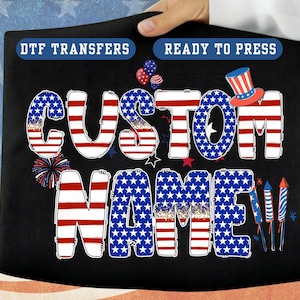 May include: Black DTF transfer with the words "CUSTOM NAME" in red, white, and blue, with a patriotic theme. Includes a top hat, fireworks, and balloons. Ready to press.