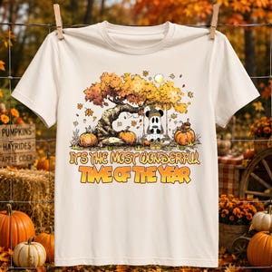 May include: A cream-colored short-sleeve t-shirt featuring a graphic of a ghost sitting on a swing under an autumn tree with pumpkins. The text reads, "IT'S THE MOST WONDERFUL TIME OF THE YEAR" in orange and yellow letters with a dark outline.