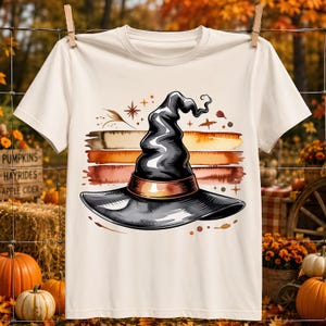May include: A cream-colored short-sleeve t-shirt featuring a graphic print of a black witch hat centered over horizontal brush strokes in shades of orange, brown, and yellow, accented with small star and leaf illustrations.