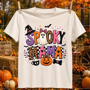 May include: A cream-colored short-sleeve t-shirt featuring a graphic print with the text SPOOKY MAMA. The design includes a ghost with a pink leopard print bow, a jack-o'-lantern with a bow, a witch hat, a coffee mug, a bone, bats, and stars.