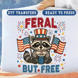 May include: A white DTF transfer with a cartoon raccoon wearing a patriotic hat and holding American flags. The text reads "FERAL BUT FREE" in red and blue. Fireworks and stars complete the design. The words "DTF TRANSFERS" and "READY TO PRESS" are at the top.