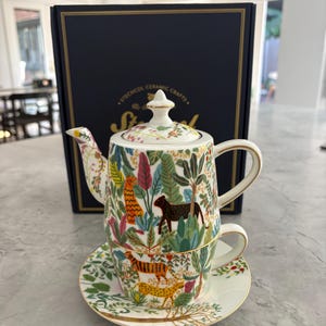 May include: A white ceramic teapot, teacup, and saucer set with a jungle animal design. The teapot and teacup are stacked, and the saucer is beneath the teacup. The set has gold trim and is in front of a navy blue box.