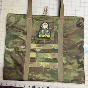 May include: Camouflage-patterned tote bag with a zipper closure and olive green straps. The bag features a patch with the word "CONCERN" and an illustration of a person. The bag has a rectangular shape and is designed for carrying items.