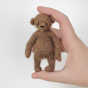 Peut inclure: Un petit ours en peluche tricot&eacute; marron avec un nez et des yeux cousus en noir. L'ours est tenu dans une main sur un fond blanc. Les bras et les jambes de l'ours sont courts et trapus, et ses oreilles sont arrondies.