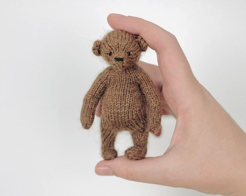 Peut inclure: Un petit ours en peluche tricot&eacute; marron avec un nez et des yeux cousus en noir. L'ours est tenu dans une main sur un fond blanc. Les bras et les jambes de l'ours sont courts et trapus, et ses oreilles sont arrondies.