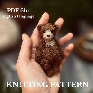 Op de afbeelding: Een kleine, bruine teddybeer met een witte borst wordt in een hand gehouden. De beer is gemaakt van een textuurrijk materiaal. Tekst op de afbeelding luidt "PDF file English language" en "KNITTING PATTERN".