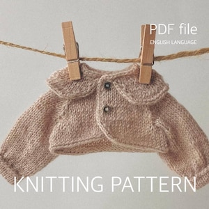 Könnte beinhalten: Eine beige gestrickte Strickjacke mit abgerundetem Kragen und zwei kleinen Knöpfen, die an einer Schnur mit zwei Holzklammern hängt. Das Bild enthält den Text PDF file, ENGLISH LANGUAGE und KNITTING PATTERN in weißer Schrift.