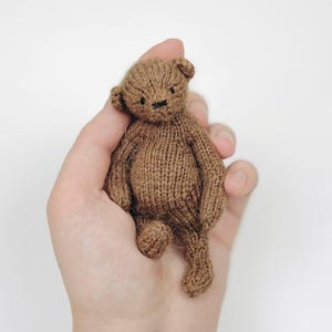 Peut inclure: Un petit ours en peluche tricot&eacute; marron avec un design d&eacute;taill&eacute;. L'ours a de petites oreilles, un nez cousu et un design simple et classique. L'ours est tenu dans une main sur fond blanc. L'ours mesure environ 10 cm de haut.