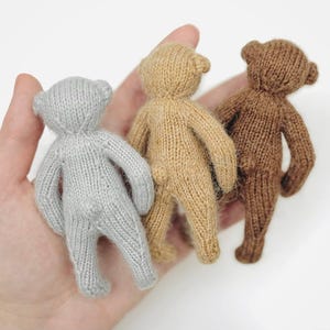 Peut inclure: Trois petits ours en peluche tricot&eacute;s, pr&eacute;sent&eacute;s dans une main. Les ours sont de couleurs grise, beige et marron. Chaque ours a un corps arrondi, de petits bras et jambes, et une petite queue. Les ours sont faits &agrave; la main et ont un aspect doux et textur&eacute;.