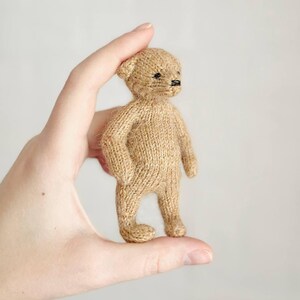 Peut inclure: Un petit ours en peluche tricot&eacute; de couleur beige avec des yeux et un nez noirs. L'ours est tenu dans une main, mettant en valeur sa taille et ses d&eacute;tails. Il a la forme classique d'un ours en peluche, avec des bras, des jambes et des oreilles.