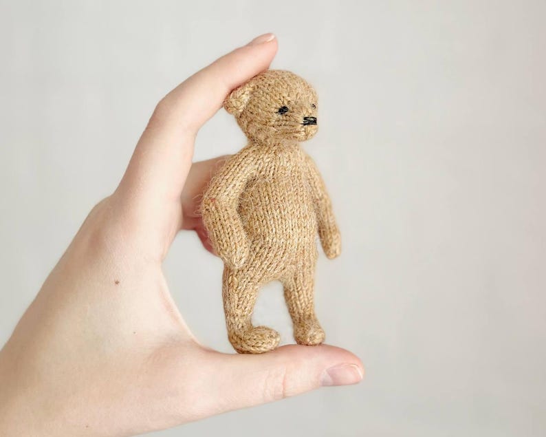 Peut inclure: Un petit ours en peluche tricot&eacute; de couleur beige avec des yeux et un nez noirs. L'ours est tenu dans une main, mettant en valeur sa taille et ses d&eacute;tails. Il a la forme classique d'un ours en peluche, avec des bras, des jambes et des oreilles.