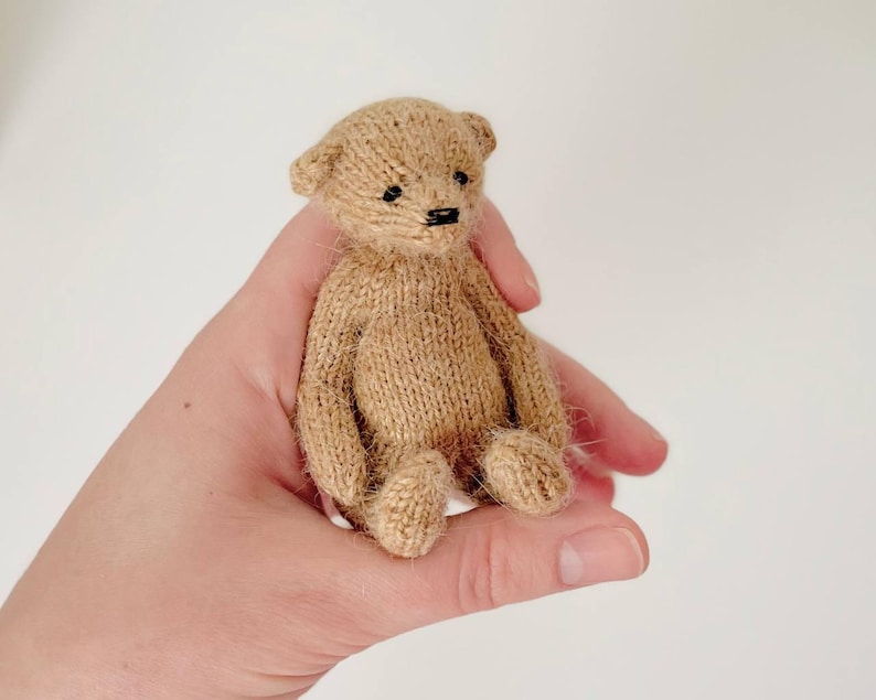 Peut inclure: Un petit ours en peluche tricot&eacute; beige avec des yeux noirs et un nez cousu noir. L'ours est assis droit, les bras et les jambes tendus. L'ours est tenu dans la main d'une personne.