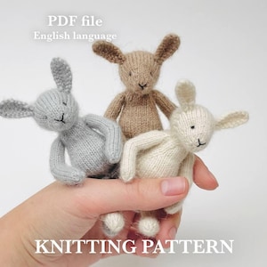 Puede incluir: Tres pequeños juguetes de conejito tejidos en gris, marrón y crema se sostienen en una mano. Los conejitos tienen orejas largas y rasgos faciales cosidos en negro. La imagen incluye el texto "PDF file English language" y "KNITTING PATTERN".