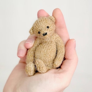 Peut inclure: Un petit ours en peluche tricot&eacute;, dans des tons beige et marron. L'ours a de petits yeux noirs et un nez cousu noir. Il est tenu dans une main ouverte, soulignant sa taille et sa texture. Les membres de l'ours sont cousus et articul&eacute;s.