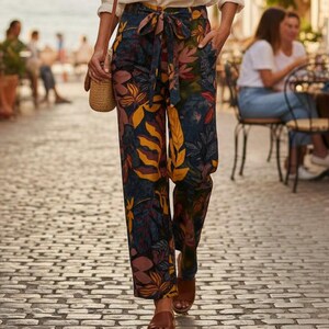 Puede incluir: Una blusa blanca de manga larga con escote en pico, combinada con pantalones de talle alto y pernera ancha con base azul oscuro y un estampado botánico vibrante de hojas amarillas, naranjas y rosas, que incluye un cinturón de tela a juego.