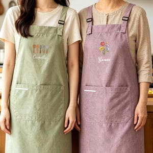 以下が含まれることがあります： 調節可能なストラップと2つのフロントポケットが付いた2枚のエプロン。セージグリーンのエプロンにはキッチン用品のグラフィックと「Camilla」という名前が、モーブカラーのエプロンにはカラフルな花束のグラフィックと白い筆記体で「Sloane」という名前がプリントされています。
