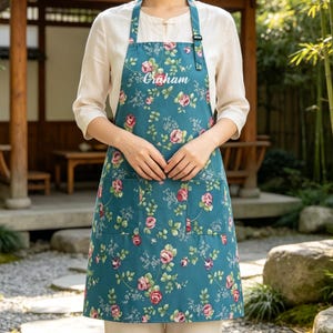 以下が含まれることがあります： ピンクのバラと緑の葉のフローラル柄が施されたティールカラーのエプロン。胸元には白い筆記体でGrahamという名前がプリントされています。エプロンには2つのフロントポケットと、黒いプラスチック製のバックルが付いた調節可能なネックストラップが付いています。