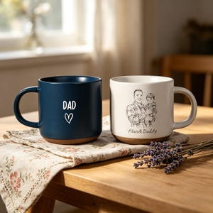 Puede incluir: Dos tazas de cerámica sobre una mesa de madera. Una es azul marino con "DAD" y un corazón. La otra es blanquecina con un boceto de un hombre y un niño, con el texto "Alan & Daddy". Ambas tienen bases marrones.