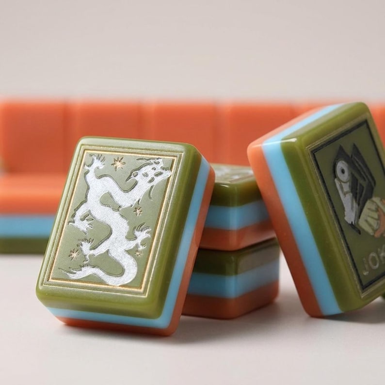 Peut inclure: Un ensemble de tuiles de mah-jong rectangulaires avec un design en couches, comprenant un dessus vert olive, un milieu bleu clair et une base orange. Une tuile pr&eacute;sente un dragon blanc grav&eacute;, une autre un motif noir et le texte JOKER.