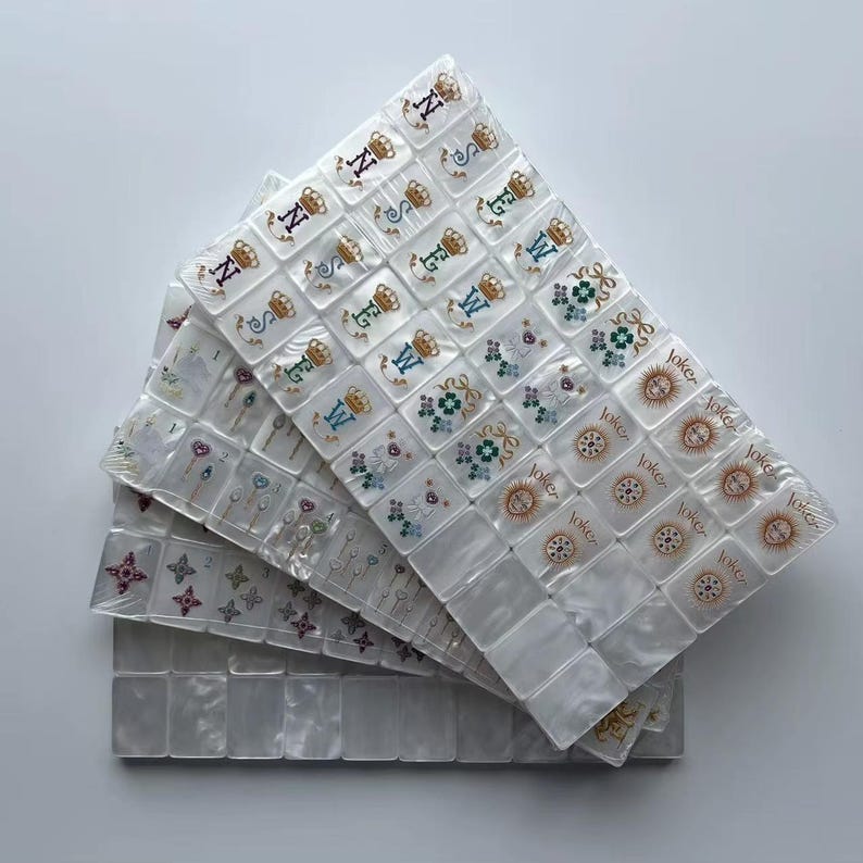 K&ouml;nnte beinhalten: Ein Set aus wei&szlig;en, perlmuttfarbenen Mahjong-Steinen, die in &uuml;berlappenden Schichten angeordnet sind. Die Steine zeigen farbige Gravuren, darunter Buchstaben mit Kronen, Blumenmuster und Joker-Steine mit dem Wort Joker in schwarzer Schrift.