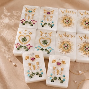 K&ouml;nnte beinhalten: Ein Set aus rechteckigen wei&szlig;en Fliesen mit detaillierten, gepr&auml;gten Designs in Gold, Gr&uuml;n, Blau und Violett. Einige Fliesen zeigen Schmetterlinge, Herzen und Kleebl&auml;tter, w&auml;hrend andere das Wort Joker mit einem Strahlenmuster tragen.