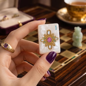 K&ouml;nnte beinhalten: Ein rechteckiger, perlmuttwei&szlig;er Mahjong-Spielstein mit der blauen Ziffer 1 und einem zentralen goldenen Filigran-Emblem, das mit einem ovalen rosa Stein, gr&uuml;nen Akzenten und kleinen klaren Kristallen verziert ist, gehalten zwischen Fingern mit dunkelviolettem Nagellack.