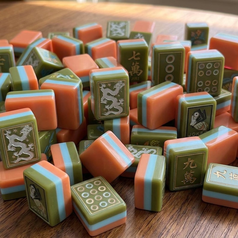 Peut inclure: Un ensemble de tuiles de Mahjong pr&eacute;sentant un design en couches avec des sections vert olive, bleu clair et orange. Les tuiles affichent divers symboles grav&eacute;s, notamment des caract&egrave;res chinois, des dragons et des ic&ocirc;nes joker, avec des points num&eacute;riques.