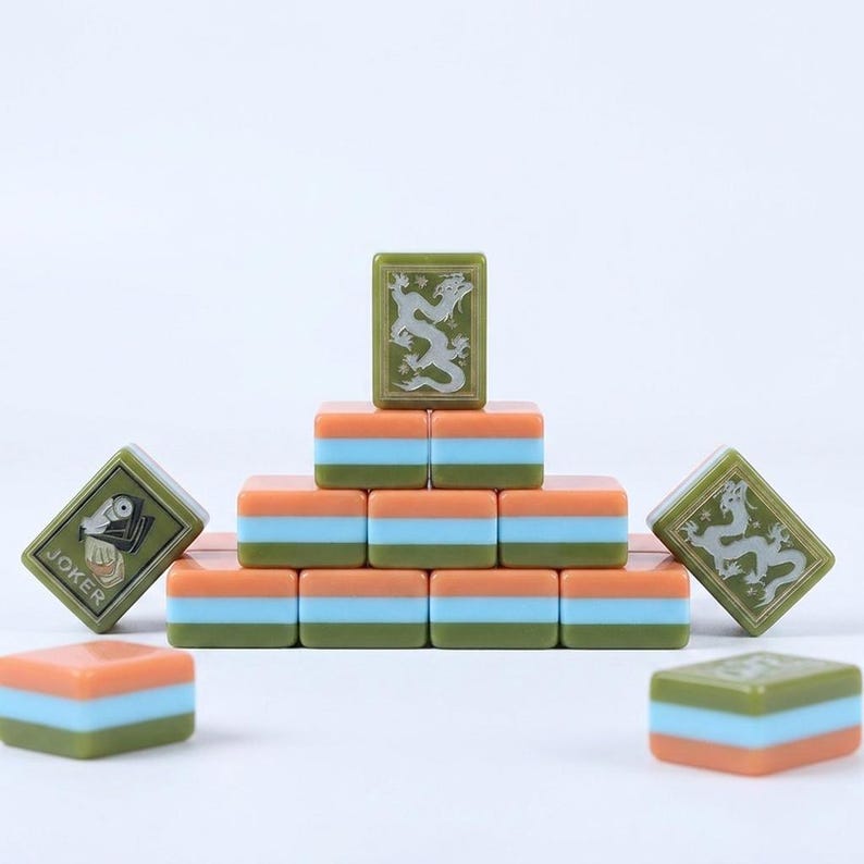 Peut inclure: Un ensemble de tuiles de mah-jong rectangulaires empil&eacute;es en pyramide. Les tuiles pr&eacute;sentent un design tricolore vert olive, bleu clair et orange. Certaines tuiles arborent un motif de dragon grav&eacute;, tandis qu'une tuile pr&eacute;sente une illustration joker.