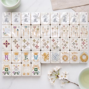 K&ouml;nnte beinhalten: Ein Set aus rechteckigen Mahjong-Spielsteinen mit perlmuttwei&szlig;er Oberfl&auml;che. Die Steine zeigen verschiedene eingravierte Motive, darunter die Zahlen eins bis neun, Zepter, Sternformen, Kronen, die Buchstaben N, S, E, W, L&ouml;wen und das Wort Joker.