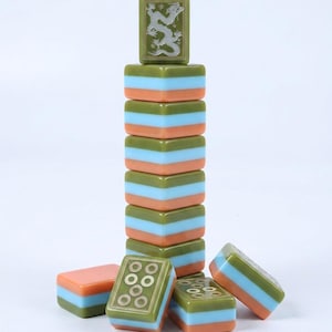 Peut inclure: Une pile de tuiles de mah-jong rectangulaires pr&eacute;sentant un design tricolore vert olive, bleu clair et orange p&ecirc;che. La tuile sup&eacute;rieure affiche un motif de dragon grav&eacute;, tandis que les autres tuiles montrent des motifs circulaires.