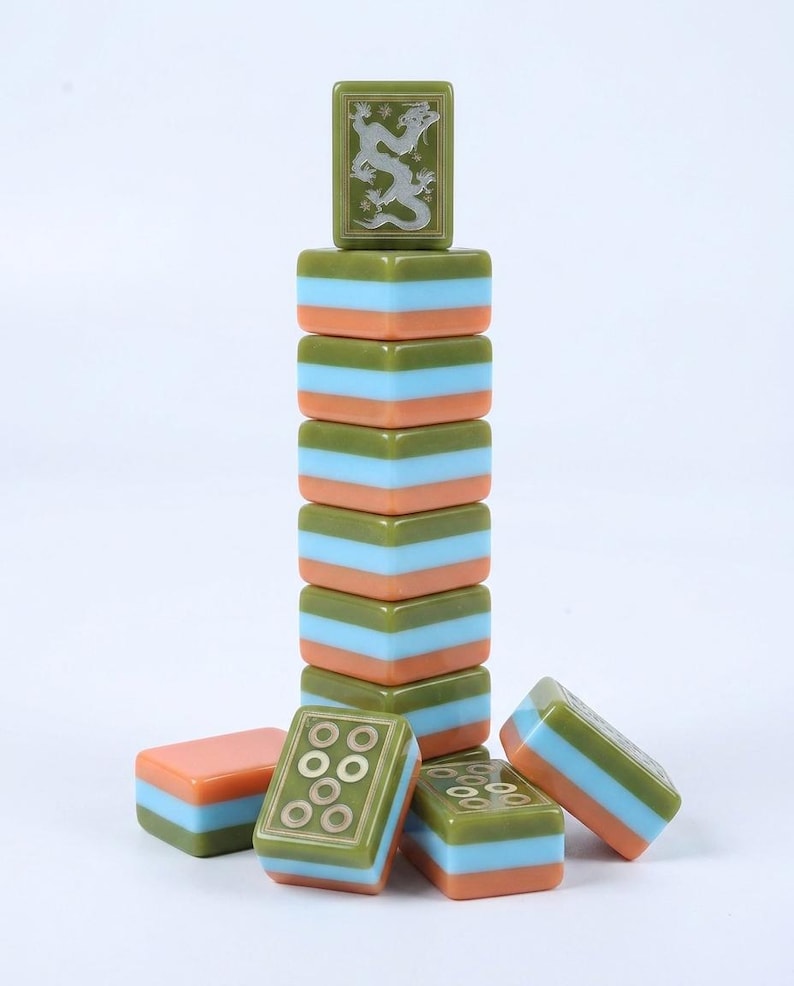 Peut inclure: Une pile de tuiles de mah-jong rectangulaires pr&eacute;sentant un design tricolore vert olive, bleu clair et orange p&ecirc;che. La tuile sup&eacute;rieure affiche un motif de dragon grav&eacute;, tandis que les autres tuiles montrent des motifs circulaires.