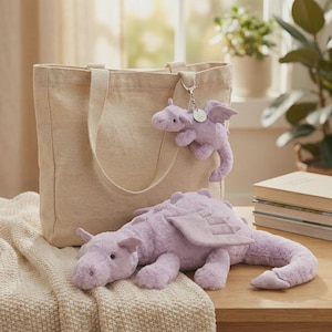 May include: A light beige tote bag with matching handles, a small lavender dragon keychain, and a large lavender plush dragon. The plush dragon is lying on a cream-colored blanket on a wooden surface.