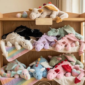 May include: A wooden shelf displays a collection of plush dragon toys in various colors. Dragons are white, black, blue, pink, purple, and green. Some have wings and decorative accents. The shelf also holds decorative eggs.