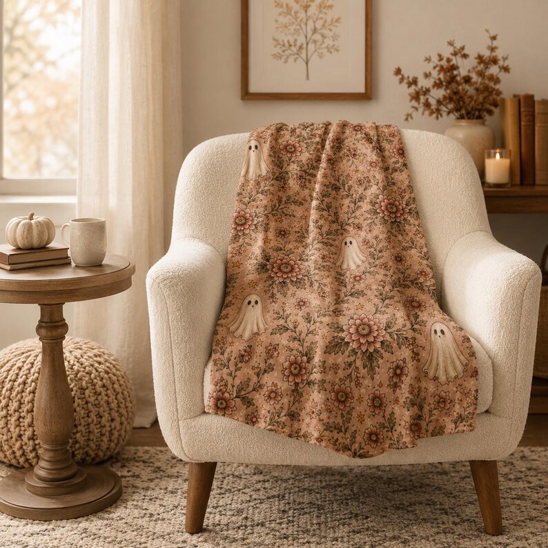 May include: A soft throw blanket featuring a repeating pattern of small white ghosts interspersed among detailed floral motifs in shades of muted orange, brown, and green. The blanket is draped over a cream-colored upholstered armchair.